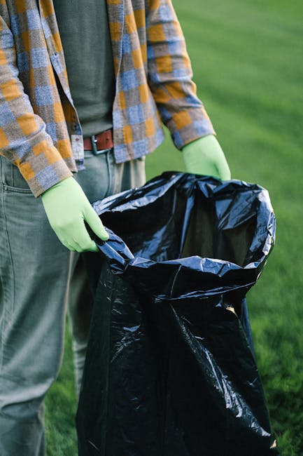 A large pile of transparent plastic garbage bags filled with mixed waste, including clothing, paper, and packaging, situated on a paved sidewalk in front of a commercial building at night. The bags ar